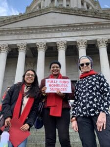 Three people smiling in front of the legislative building. The person in the middle holds a sign that says "fully fund homeless services."